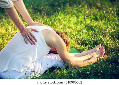 Young man and woman dressed in a white robe doing thai massage with yoga exercises sitting on the grass. Sunny summer park with green lawn in the background
