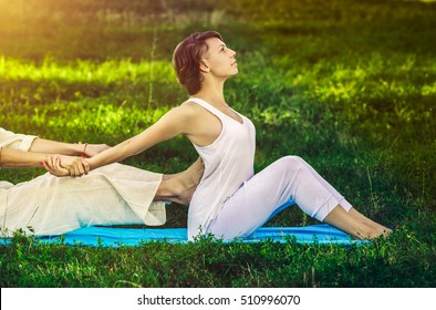 Young man and woman dressed in a white robe doing thai massage with yoga exercises sitting on the mat. Sunny summer park with green lawn in the background
