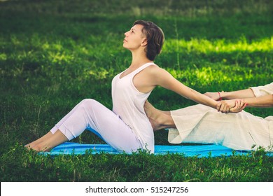 Young man and woman dressed in a white robe doing thai massage with yoga exercises sitting on the mat. Sunny summer park with green lawn in the background