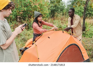 Young man and woman fixing poles and setting up tent while his friend reading online manual on phone
