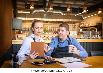 Young Man And Woman Going Through Paperwork Together In Their Restaurant. Small Family Restaurant Owners Discussing Finance Calculating Bills And Expenses Of Their Small Business.