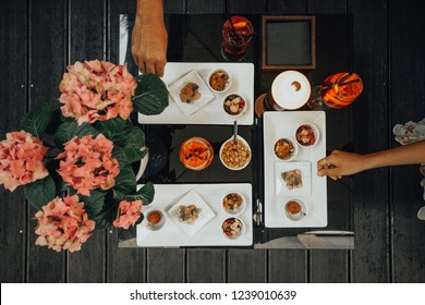 Young Man And Woman Hands Picking Snacks From Italian Aperitif With Spritz Cocktail, Drink With Aperol, Prosecco & Soda. Beautiful Dark Wooden Table  With Rose Flower Decoration Top View.