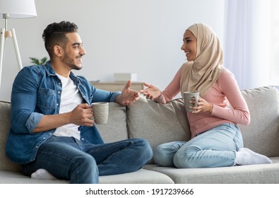 Young man and woman in hijab drinking tea and talking at home. Beautiful muslim loving couple drinking coffee while sitting on sofa at living room, spending weekend together, copy space