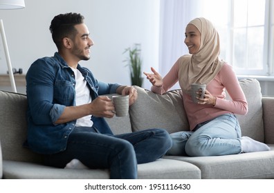 Young man and woman in hijab drinking coffee and having conversation at home. Beautiful muslim loving couple drinking tea while relaxing on sofa at living room, spending weekend together, copy space - Powered by Shutterstock