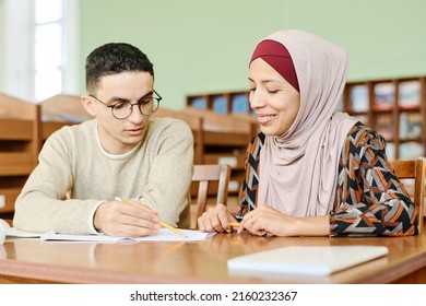 Young man and woman in hijab sitting at desk in classroom having lesson for immigrants working on writing task together - Shutterstock ID 2160232367