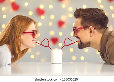Young man and woman leaning over table at home or in cafe and drinking from one cup through heart-shaped straws. Cute and funny couple moments. Having fun on Saint Valentine's Day. Sharing happiness