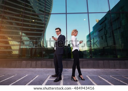 Young man and woman proud CEO are checking e-mail on smart phones, while are standing outside company where they work. Two proud economist are reading financial news via cellphone during work break