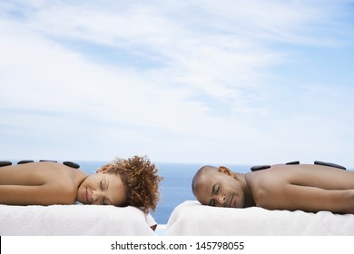 Young man and woman receiving hot stone therapy at spa with ocean in background