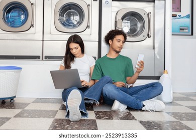 A young man and woman relax on the floor of a laundromat, engaging in different activities. The man looks at his smartphone while the woman types on her laptop, both appearing focused and comfortable. - Powered by Shutterstock