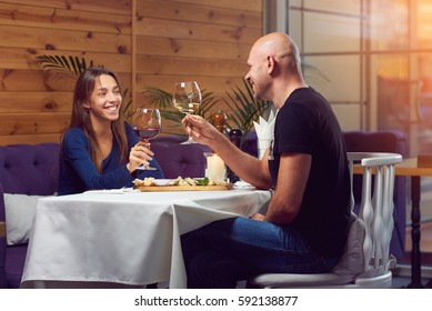 A young man and woman in the restaurant having dinner. Looking at each other and smiling while clink glasses of red and white wine  in sun light