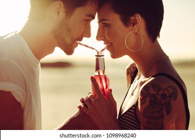 Young Man And Woman Sharing A Beverage Outdoors. Couple Drinking Soft Drink With Straws From Same Bottle.