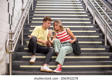 A Young Man And Woman Sitting On The Stairs Of A Subway Station. The Girl Shows The Boy Something On Her Cell Phone