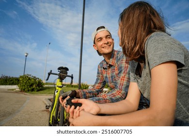 Young man and woman smiling and chatting. The man wears a plaid shirt, a cap, while the woman is in gray t-shirt. A bicycle is visible nearby against a bright sky.                                 - Powered by Shutterstock