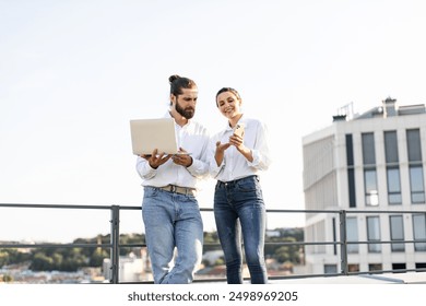 Young man and woman standing outdoors discussing work on laptop and smartphone. Casual attire, urban setting, teamwork and collaboration. Modern business concept. - Powered by Shutterstock