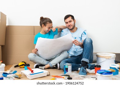 Young Man And Woman Together Planning Their Home Renovation. Cardboard Boxes, Painting Tools And Materials On Floor. House Remodeling And Interior Renovation. People Looking At Blueprint At Home.