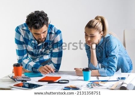 Young man and woman together working at design project. Creative teamwork at workspace with construction blueprint and color swatches. People standing near desk and discussing in architecture studio