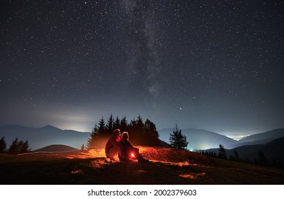 Young Man And Woman Travelers Sitting On Grassy Hill Under Night Sky With Stars. Couple Hikers Looking At Majestic Night Starry Sky And Hugging. Concept Of Hiking, Night Camping And Relationships.