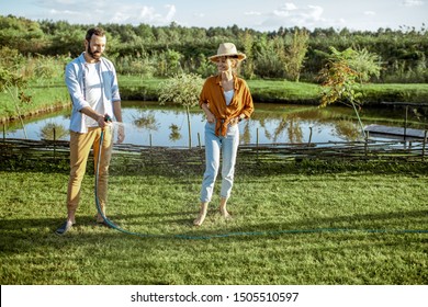 Young man and woman watering green lawn, taking care of the beautiful backyard with lake on the background
