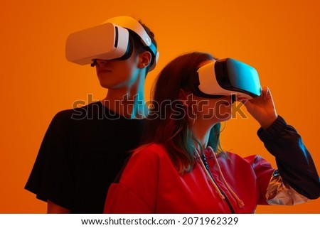 Young man and woman wearing headsets of virtual reality standing on orange background looking away