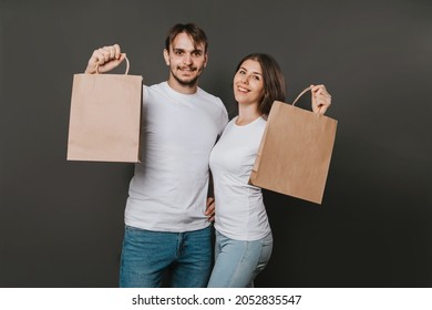 Young Man And Woman In White Blank T-shirts With Paper Bags In Their Hands On A Solid Gray Background. Studio Photo. Mock-up