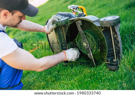 Young man worker in a special suit cleans the electric lawn mower from the grass