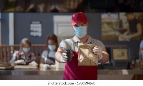 Young Man Worker Wearing Face Mask And Gloves Giving Take Away Meal To Customer. Restaurant Staff Working Only With Take Away Orders During Corona Virus Outbreak. Small Business During Pandemic