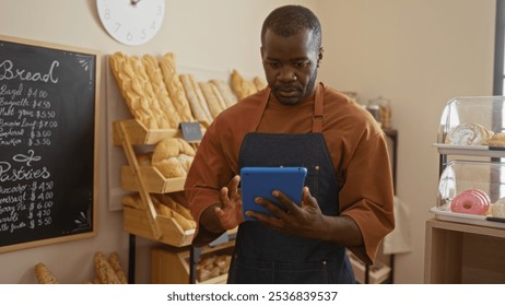 Young man working in a bakery shop, wearing an apron and using a tablet while surrounded by shelves of bread and pastries in an indoor setting - Powered by Shutterstock