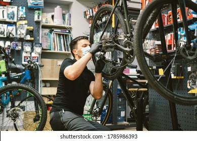 Young Man Working In A Biking Repair Shop Under Quarantine And Pandemic Coronavirus In A Protective Mask On The Face. Bicycle Mechanic Repair Bike, Cycle Workshop In Medical Mask Covid 19