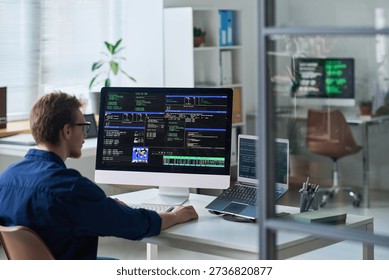Young man working at computer workstation with multiple monitors displaying programming code in office setting, focusing on software development tasks