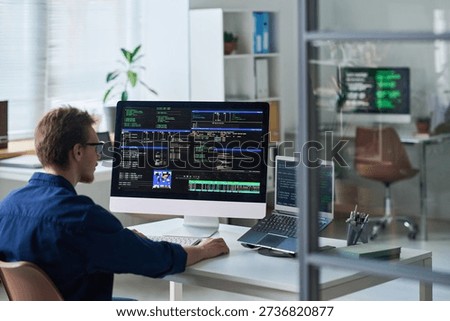 Young man working at computer workstation with multiple monitors displaying programming code in office setting, focusing on software development tasks