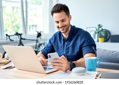 Young man working at home with laptop messaging on smartphone