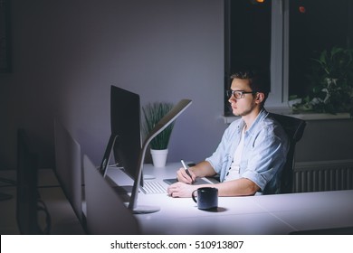 Young Man Working On Computer At Night In Dark Office. The Designer Works In The Later Time. A Young Man Sits At The Computer