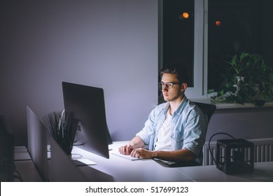 Young Man Working On Computer At Night In Dark Office. The Designer Works In The Later Time. A Young Man Sits At The Computer