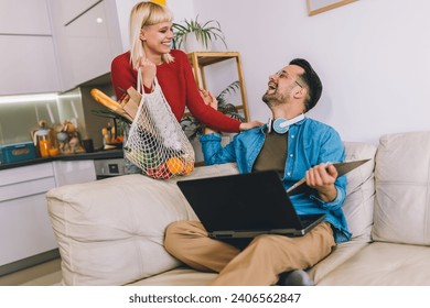 Young man is working on his laptop while wearing headphones and sitting on a comfortable couch. His wife has just arrived home from grocery shopping, holding a bag of fresh produce.