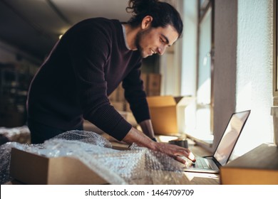 Young man working on laptop with parcel on the side. Drop shipping business owner updating the delivery status of the shipment.