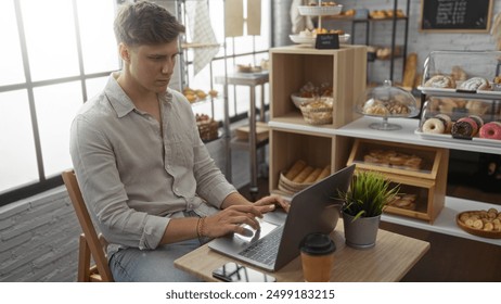 Young man working on a laptop in a bakery, surrounded by various baked goods with a cup of coffee on the table - Powered by Shutterstock