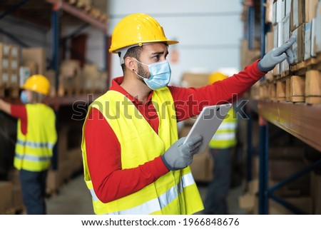 Young man working in warehouse doing inventory using digital tablet and loading delivery boxes plan while wearing face mask during corona virus pandemic - Logistic and industry concept