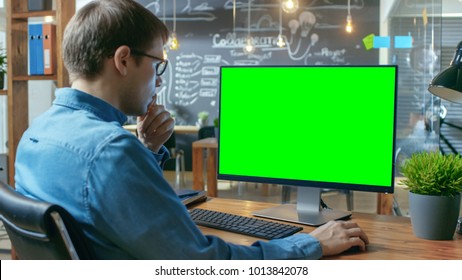 Young Man Works At His Desk On The Personal Computer With Mock-up Green Screen. In The Background His Colleague Works In The Creative Office.