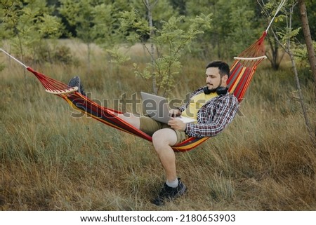 A young man works with his laptop outdoors, lounging in a hammock outdoors. A happy man lounging on a hammock in the woods, he is in a reclining position, he is wearing shorts and a yellow T-shirt.