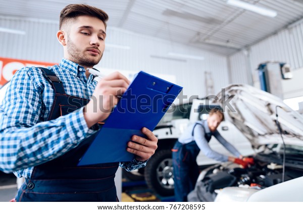 A\
young man works at a service station. The mechanic is engaged in\
repairing the car. A young mechanic is in charge of working\
records. His partner digs under the hood of the\
car.