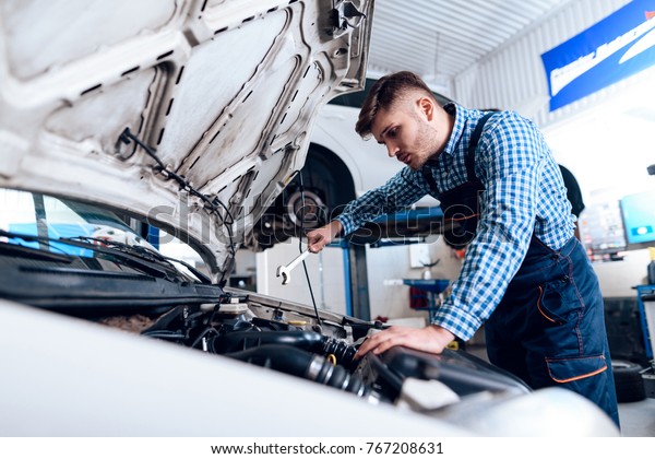 A young man works at a service station. The mechanic\
is engaged in repairing the car. Young mechanic dig under the hood\
of the car.