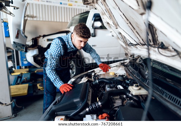 A young man works at a service station. The mechanic\
is engaged in repairing the car. Young mechanic dig under the hood\
of the car.