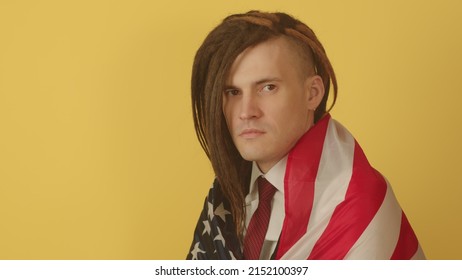 Young man wrapped in American flag on yellow background. Serious male looking at camera, posing in studio. Concept of Flag Day, national holiday