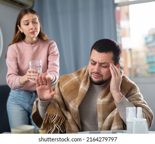 Young man wrapped in blanket sitting at table at home with cold symptoms suffering from headache, refusing with gesture of water served by worried caring wife - Shutterstock ID 2164279797