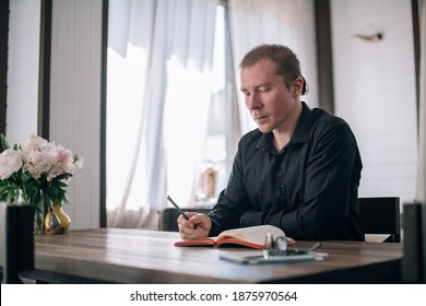 A young man writes in a notebook at the table. Journalist, writer makes notes, works on the text at the workplace in a bright room in the house - Shutterstock ID 1875970564