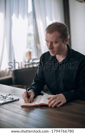 A young man writes in a notebook at the table. Journalist, writer makes notes, works on the text at the workplace in a bright room in the house