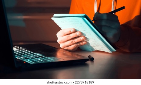 A young man writes notes in a notebook in front of a laptop, a student and a freelancer makes financial calculations, planning, studies at night at home online.