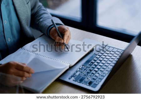 A young man writing notes by hand at a desk while using a laptop in a modern office setting