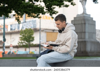 Young man writing notes in notebook with headphones in public park. Freelance 