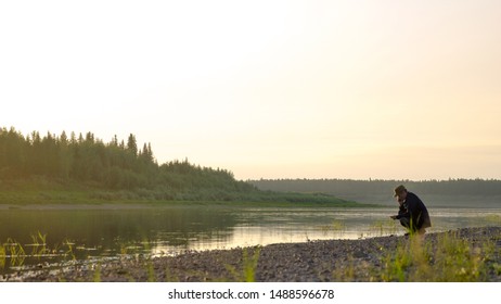 The Young Man Yakut Squatting Sklona Head Talking On The Mobile Phone On The Shore Of The Wild North Of The Vilyui River In The Forest.